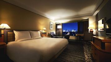 A hotel room with a large bed, two bedside tables with lamps, a desk with a chair, and a view of the city at night.
