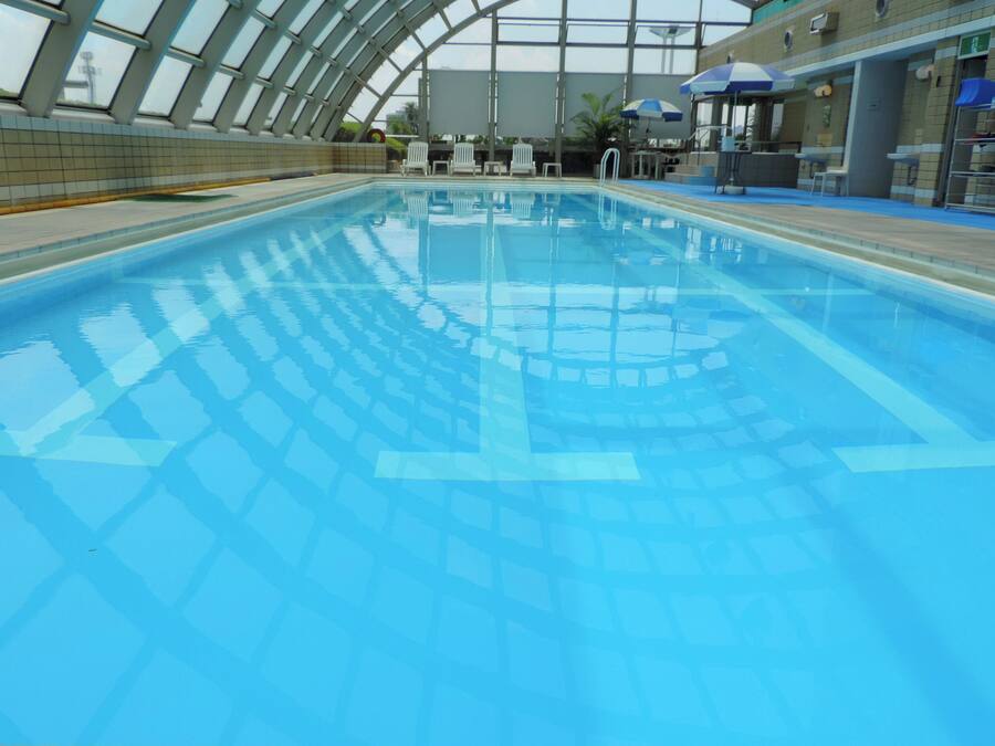 A large indoor swimming pool with clear blue water, surrounded by a tiled wall and a glass ceiling.