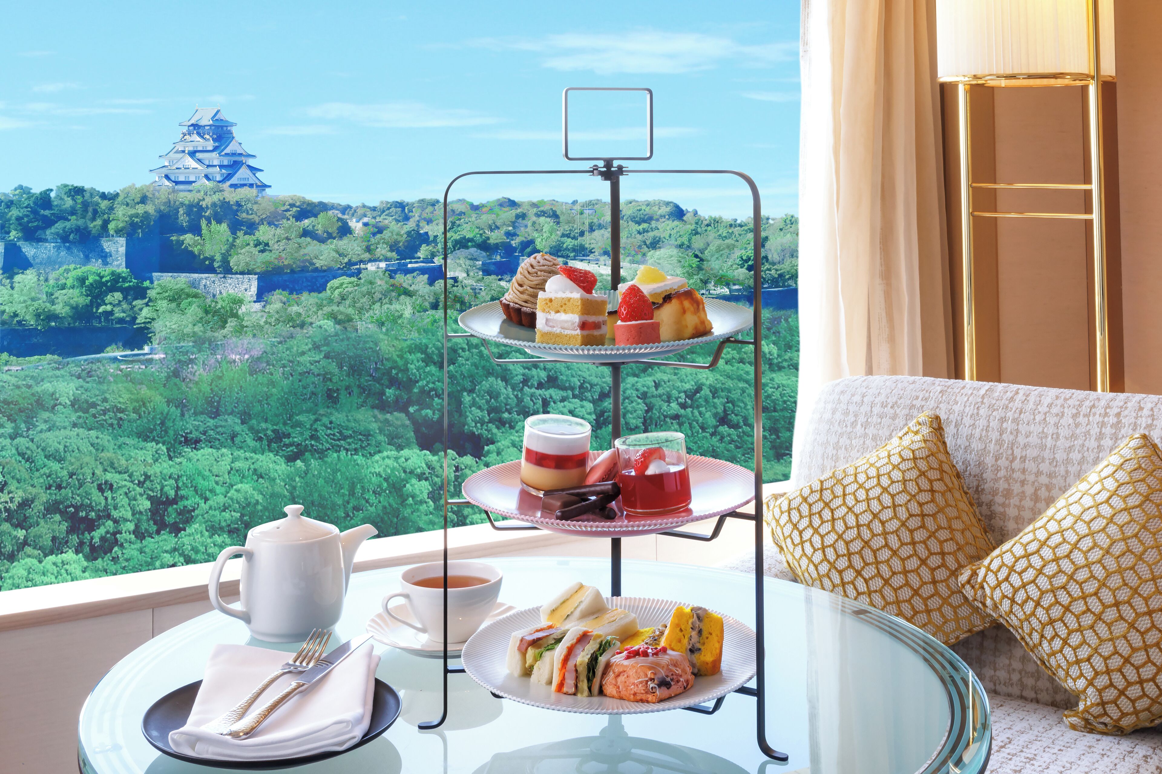 A three-tiered dessert stand with various pastries and a teapot on a table by a window overlooking a castle and lush greenery.