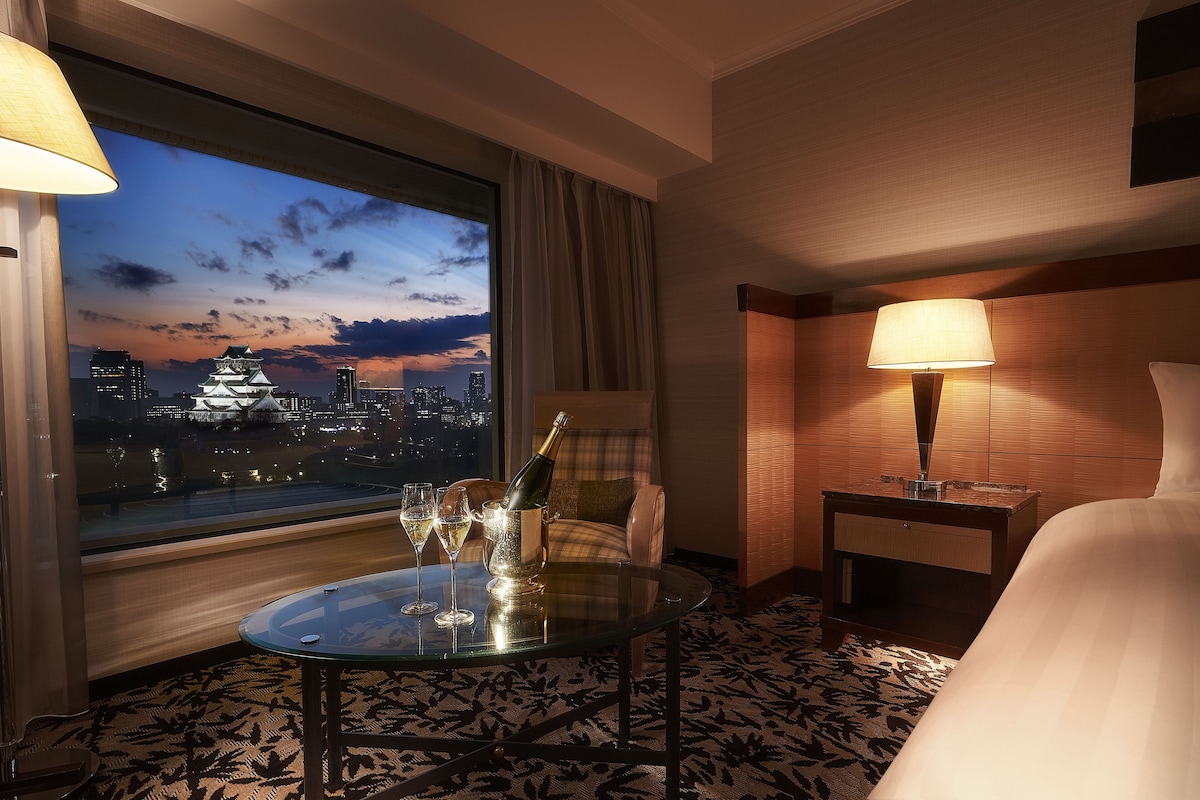 A hotel room with a large window offering a city view at dusk, a glass table with champagne and glasses, a chair, and a bedside lamp.