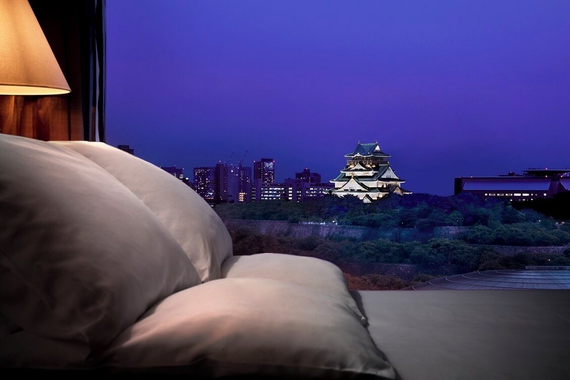 A bed with pillows, a lamp, and a view of a castle-like building at dusk.