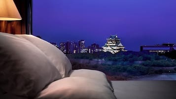 A bed with pillows, a lamp, and a view of a castle-like building at dusk.