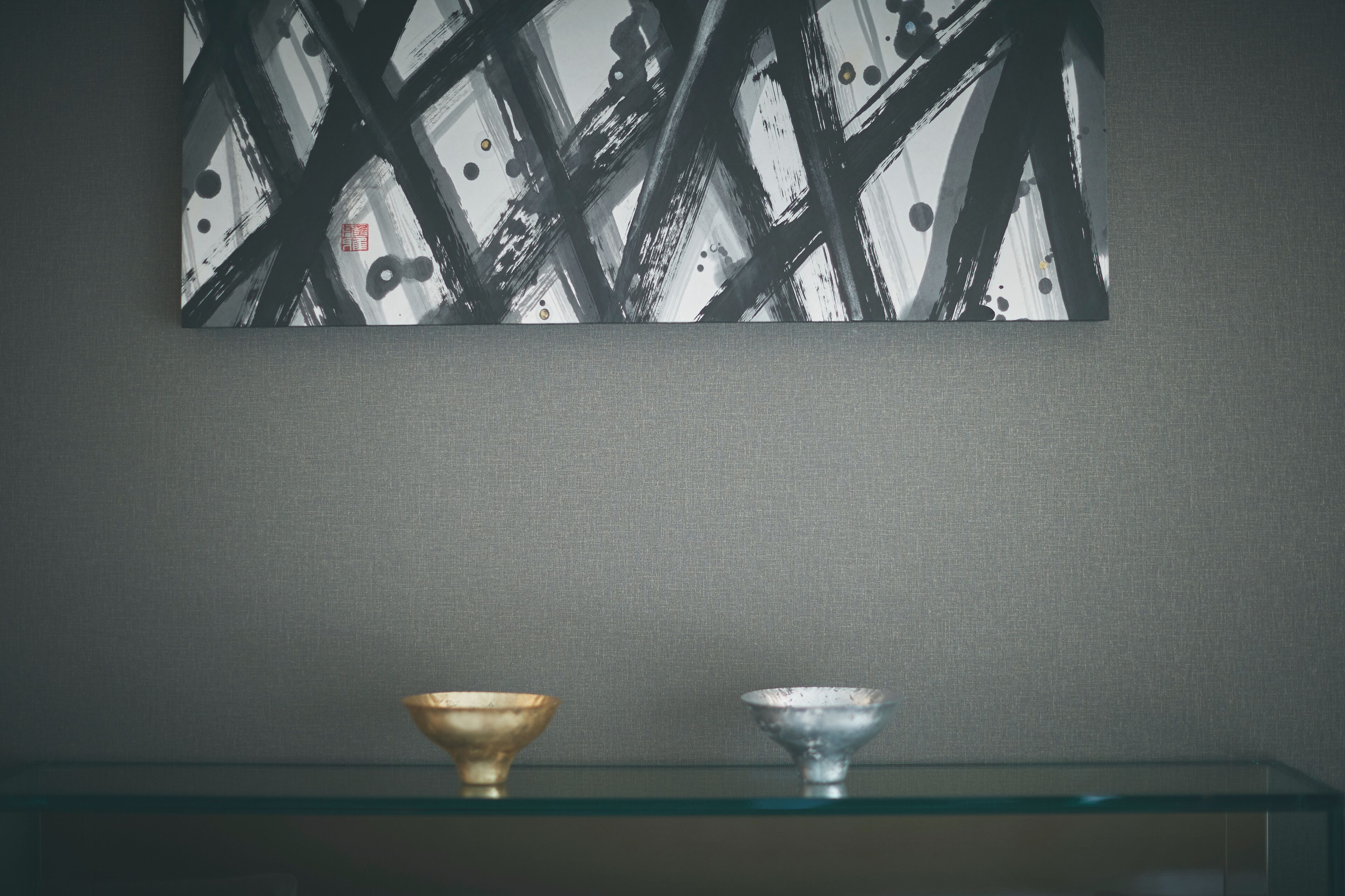 A room with a glass shelf holding two bowls, a textured grey wall, and a black and white abstract painting above.
