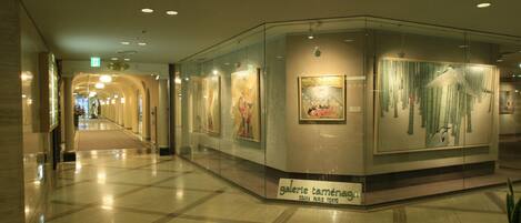 A modern hotel corridor with glass walls displaying artwork, a clear exit sign, and a polished marble floor.