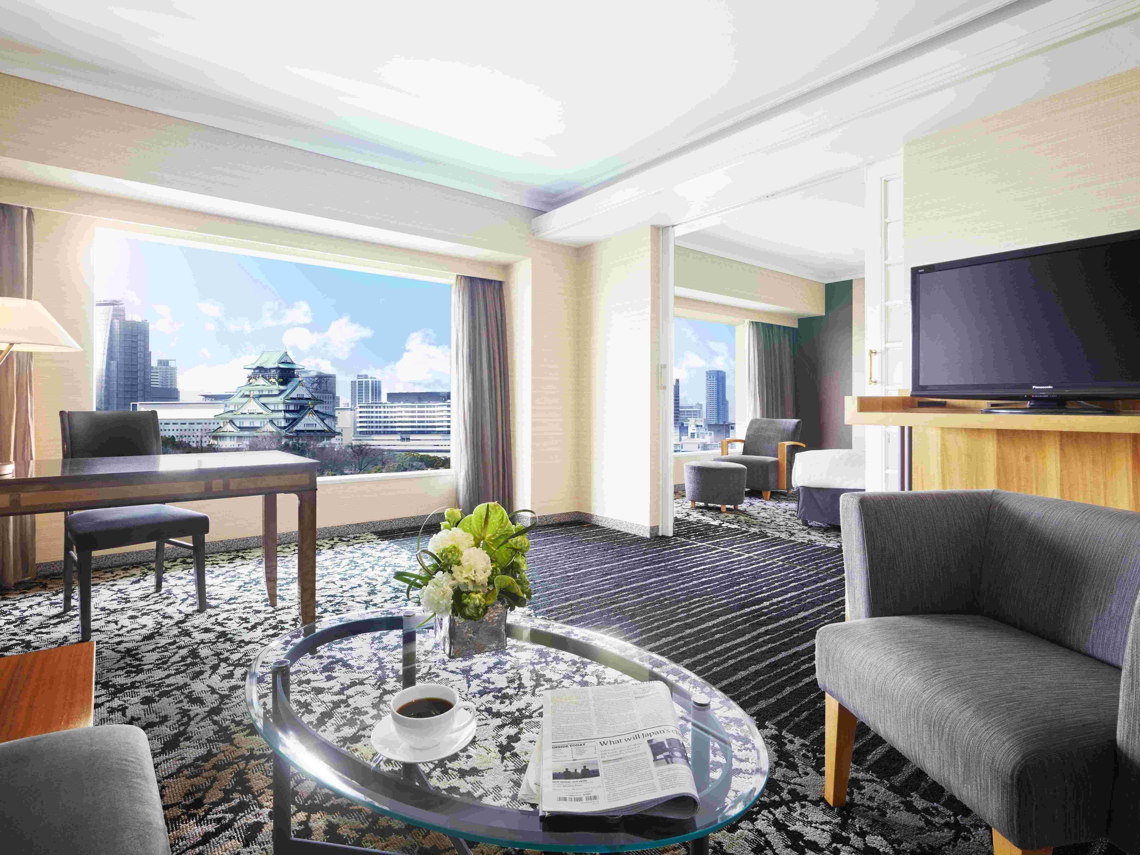 A hotel room with a city view, a flat-screen TV, a round glass coffee table with a newspaper, and a comfortable seating area.