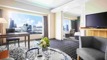 A hotel room with a city view, a flat-screen TV, a round glass coffee table with a newspaper, and a comfortable seating area.