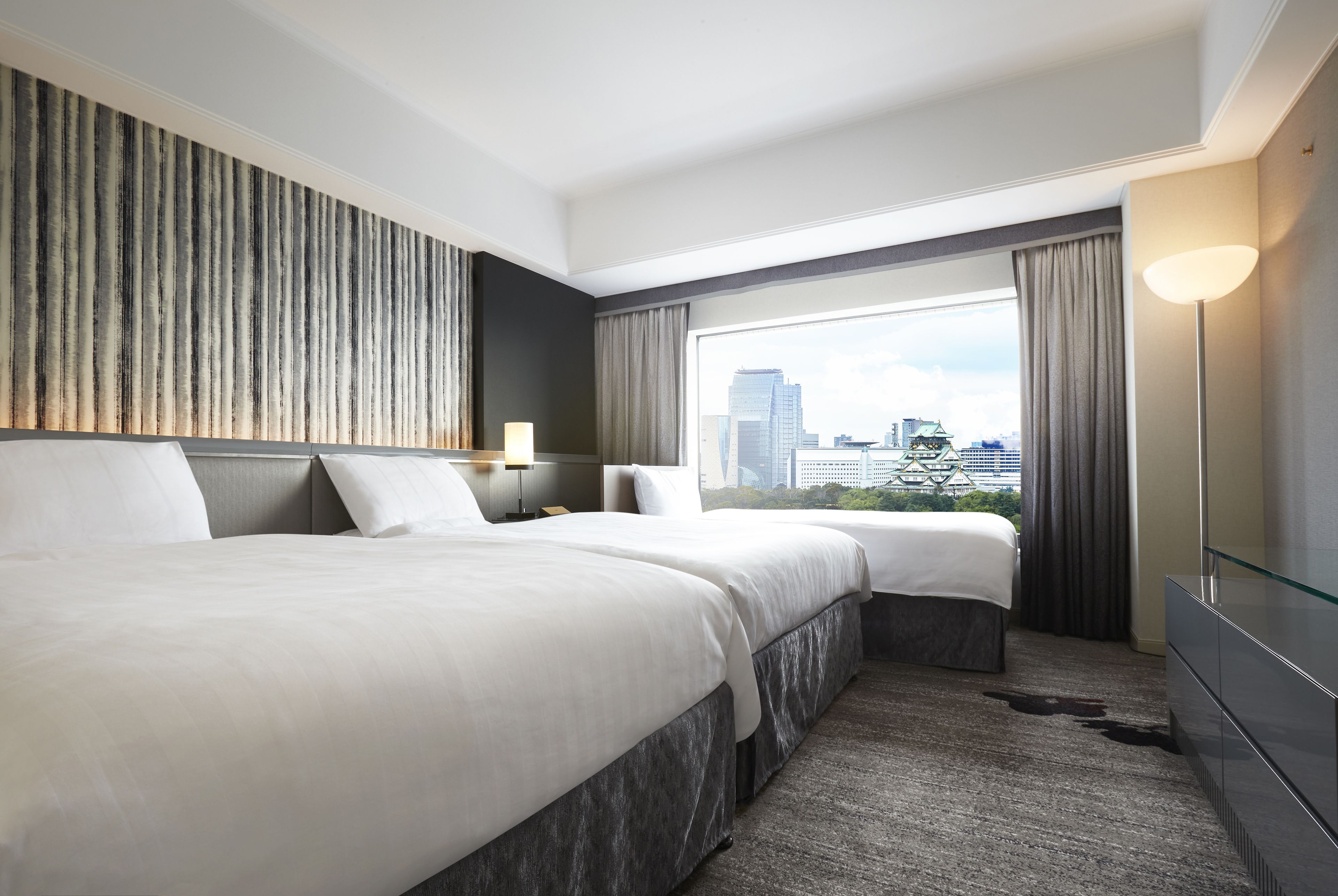 A hotel room with two beds, a large window with a city view, a modern lamp, and a flat-screen TV.