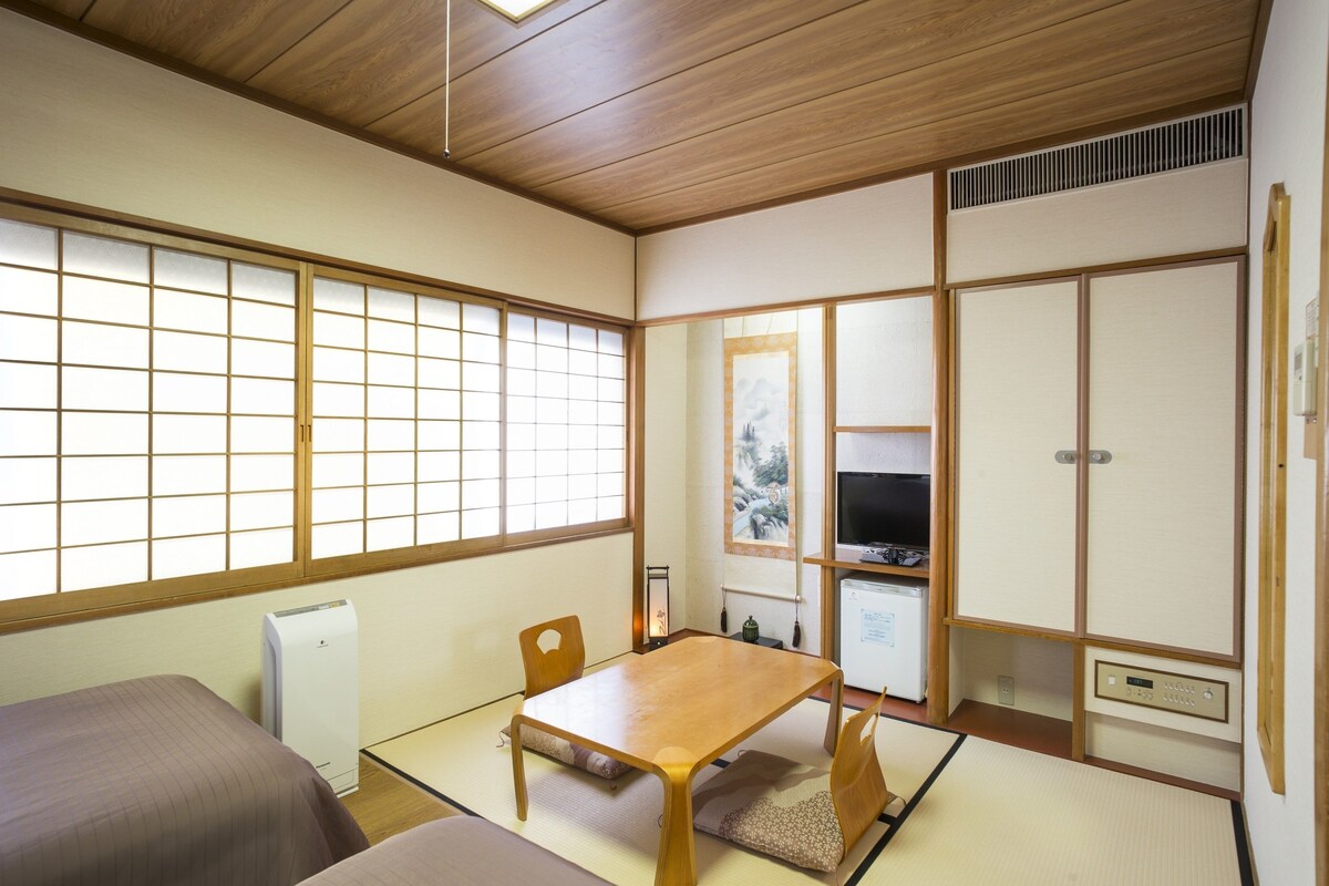 A traditional Japanese room with tatami flooring, a wooden table, two tatami beds, a television, and sliding doors.