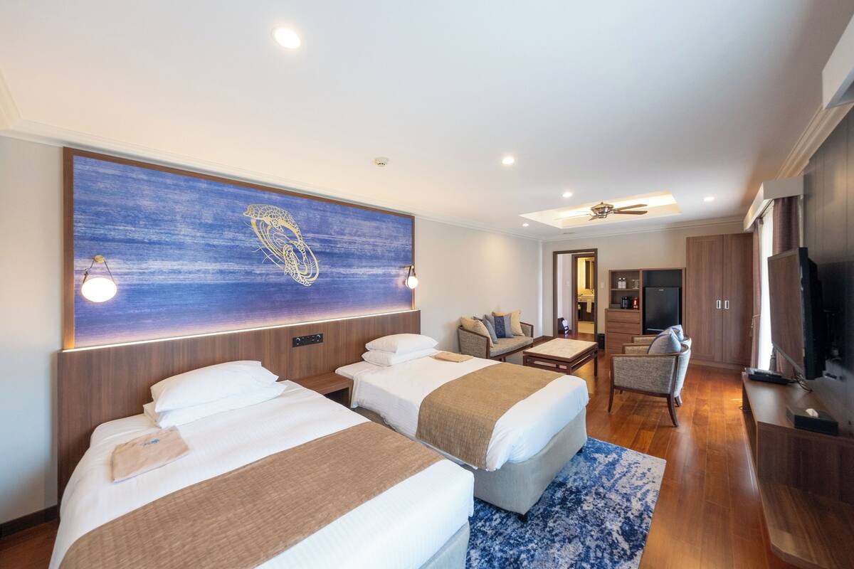 A hotel room with two beds, a large artwork on the wall, a television, and a seating area with a chair and table.