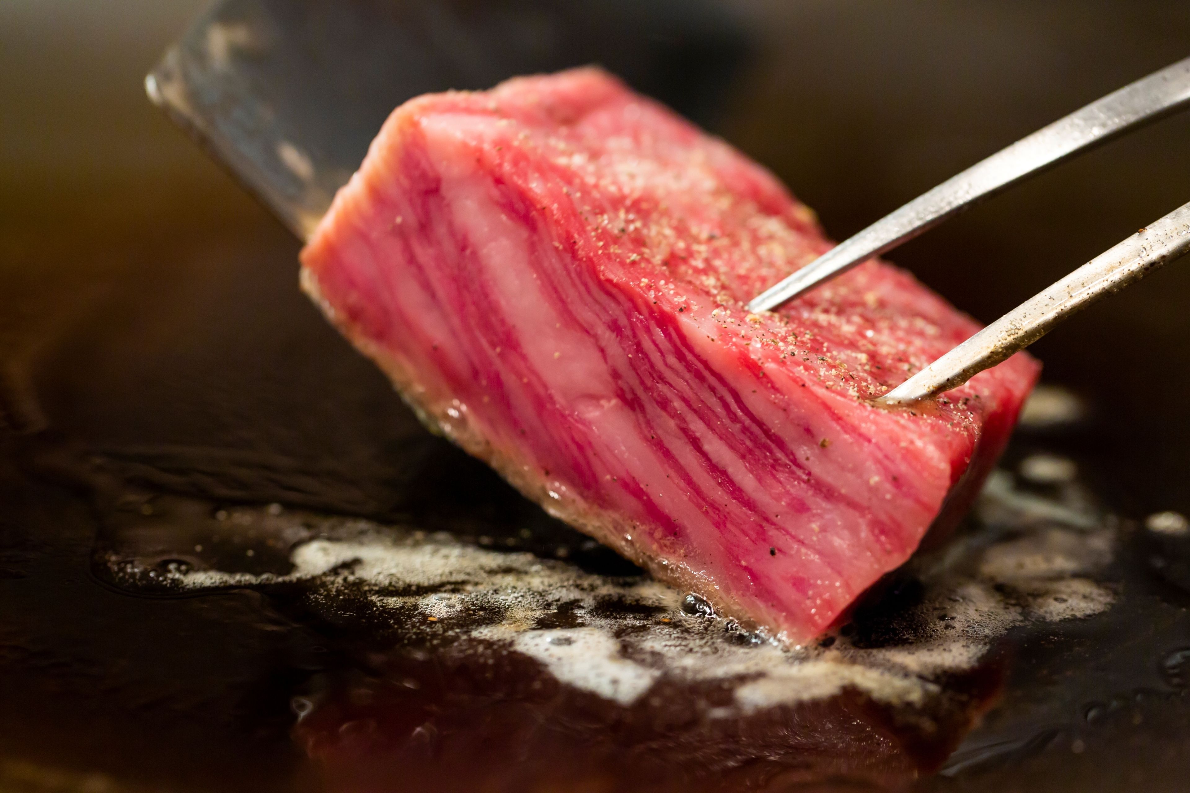 Gros plan sur un morceau de viande en train de griller, maintenu à l’aide de pinces.
