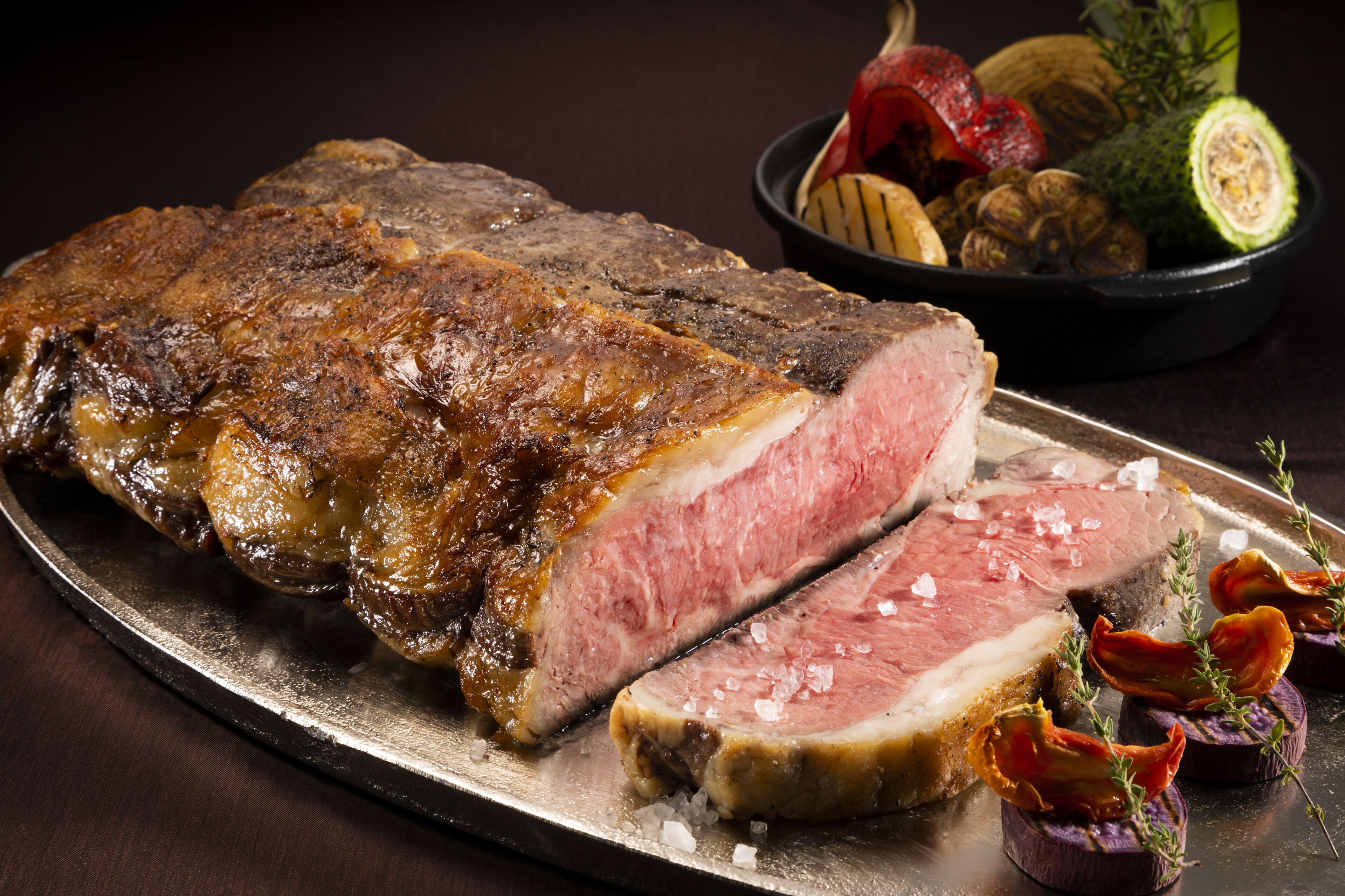 A large steak with a slice cut out, garnished with herbs and vegetables on a metal plate.