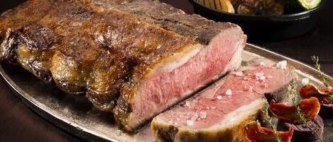 Un bistec grande con una porción retirada, adornado con hierbas y verduras sobre un plato de metal.