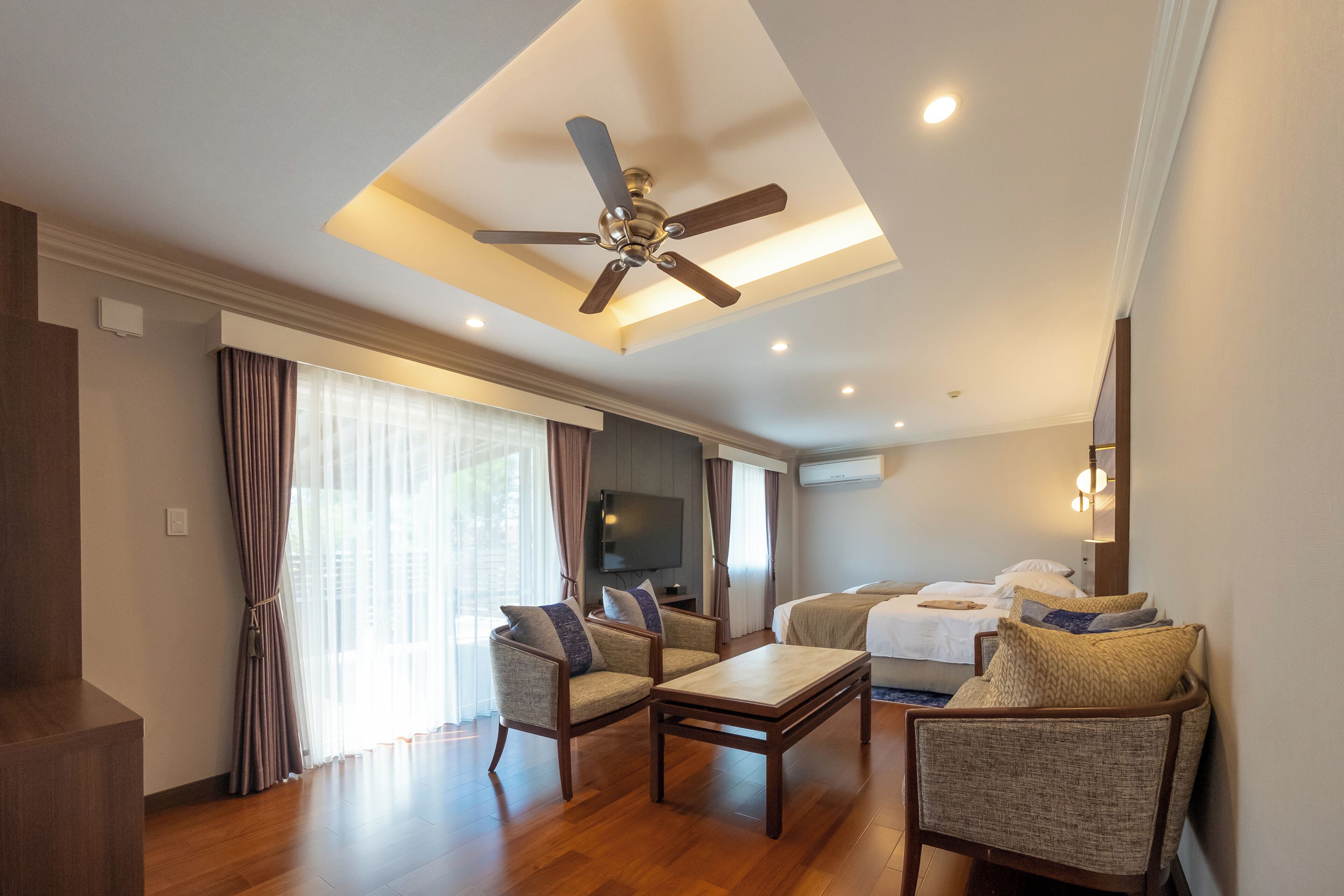 Una habitación de hotel moderna con ventilador de techo, piso de madera, televisor de pantalla plana, cama con ropa blanca y una zona de estar con dos sillones y una mesa de centro.