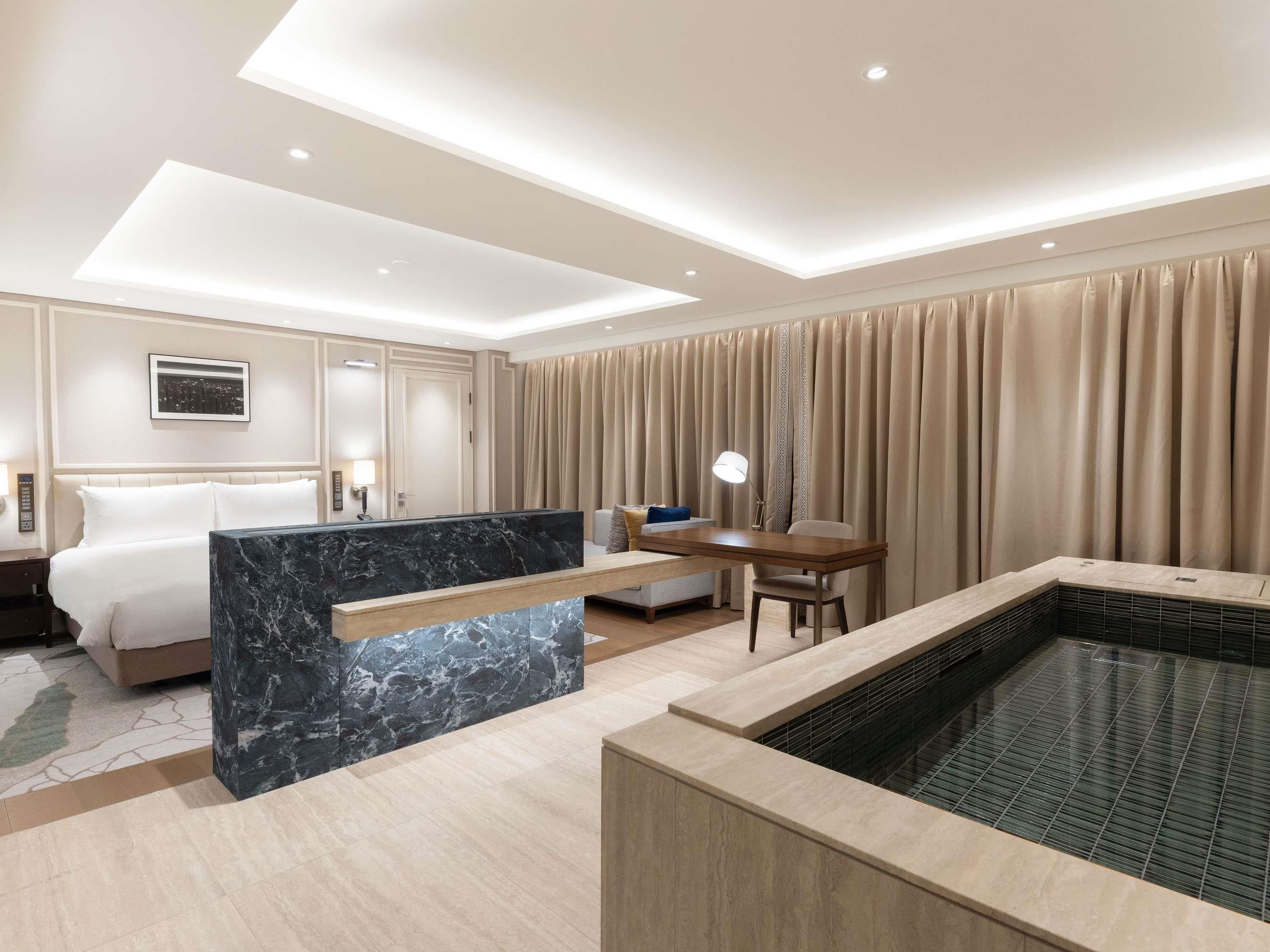 A hotel room with a bed, a desk, a chair, a marble accent wall, a large window, and a bathtub.