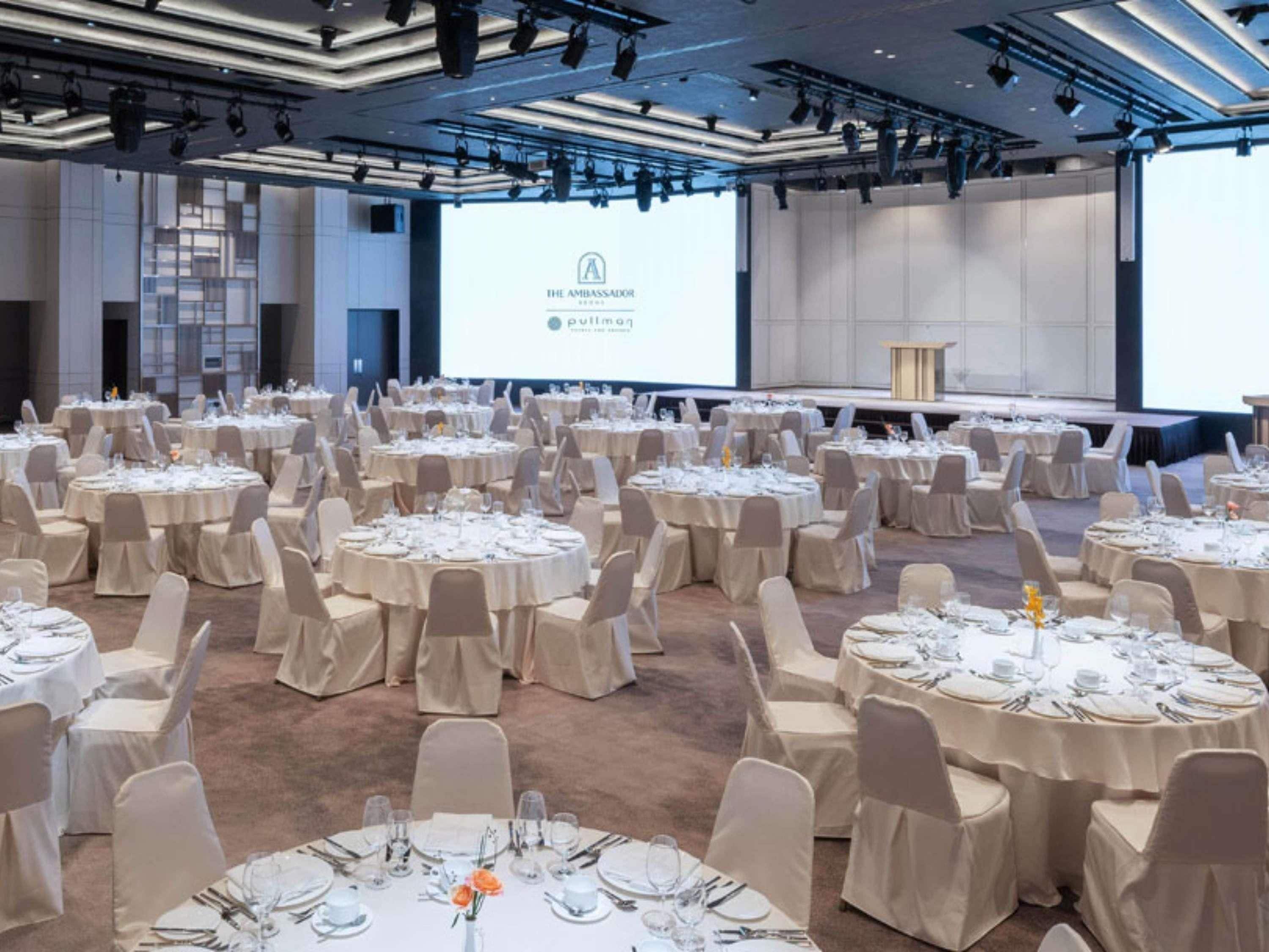 Un salón de conferencias con varias mesas redondas preparadas para un evento formal, una pantalla grande mostrando una presentación y una zona de escenario.