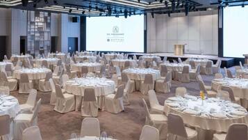 Un salón de conferencias con varias mesas redondas preparadas para un evento formal, una pantalla grande mostrando una presentación y una zona de escenario.