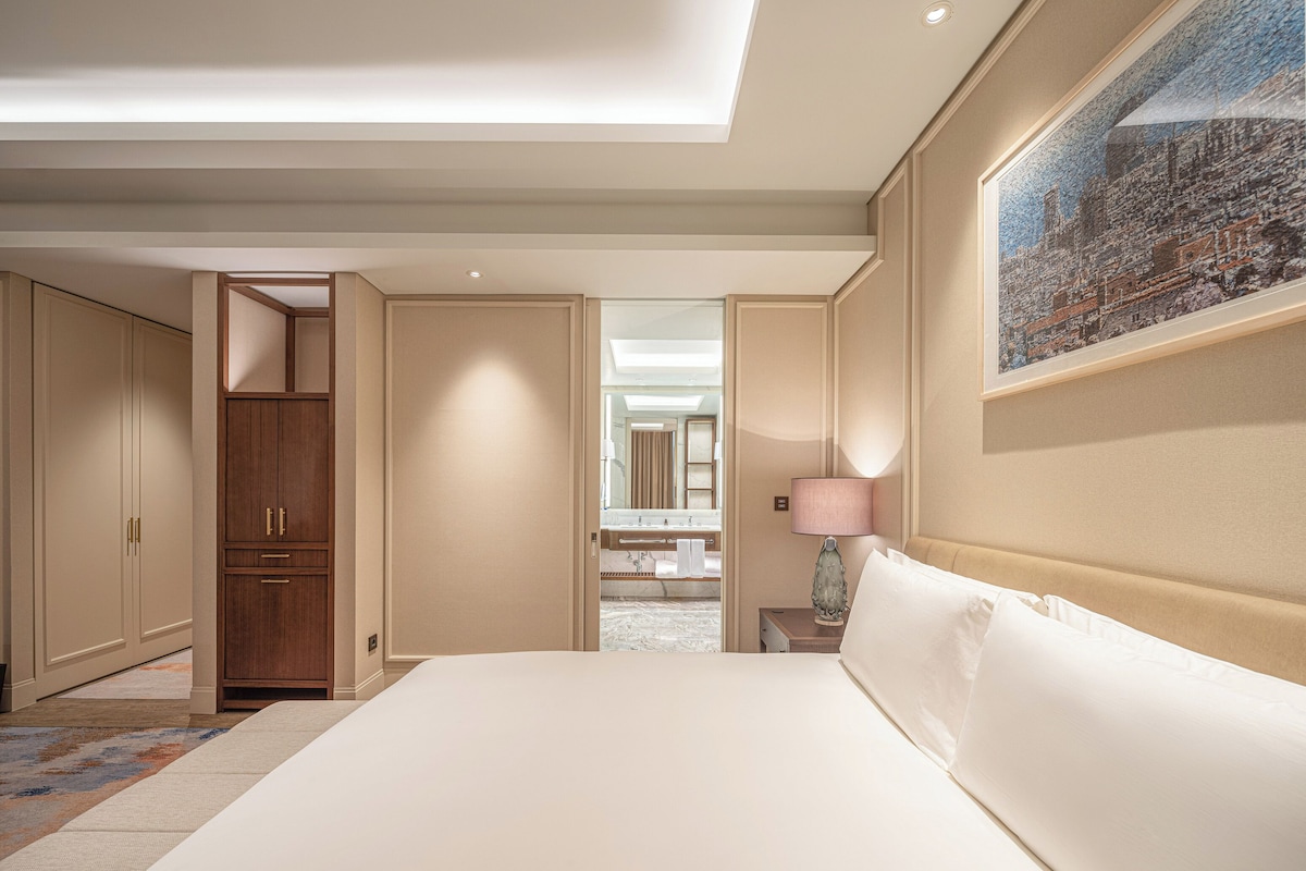 A hotel room with a large bed, a nightstand with a lamp, a wardrobe, and a bathroom visible through an open door.