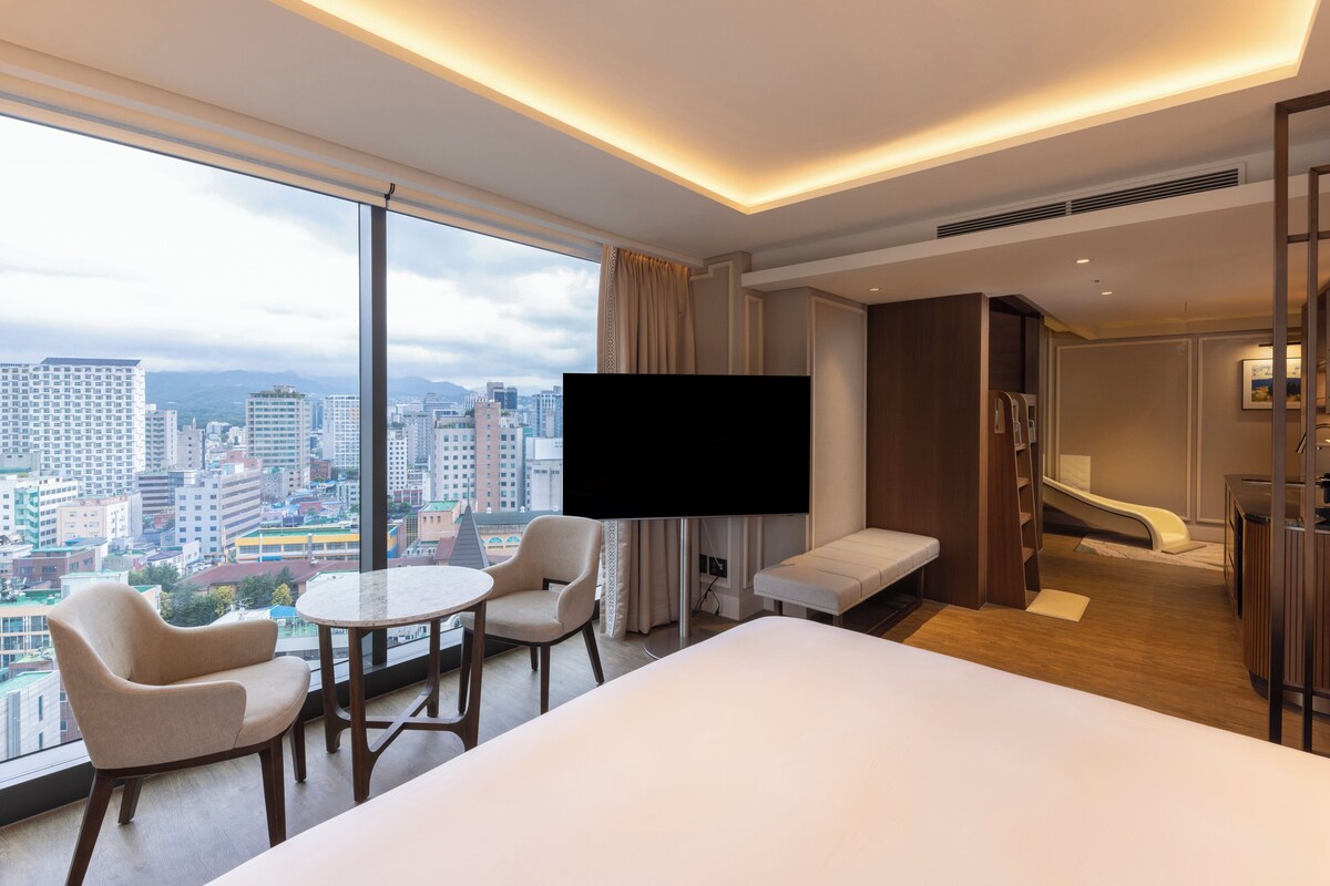A hotel room with a large bed, a TV, a seating area with chairs, a small table, and a view of the cityscape through large windows.