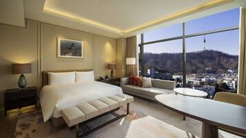 A hotel room with a bed, a sofa, a dining table, and a view of a cityscape and mountains.