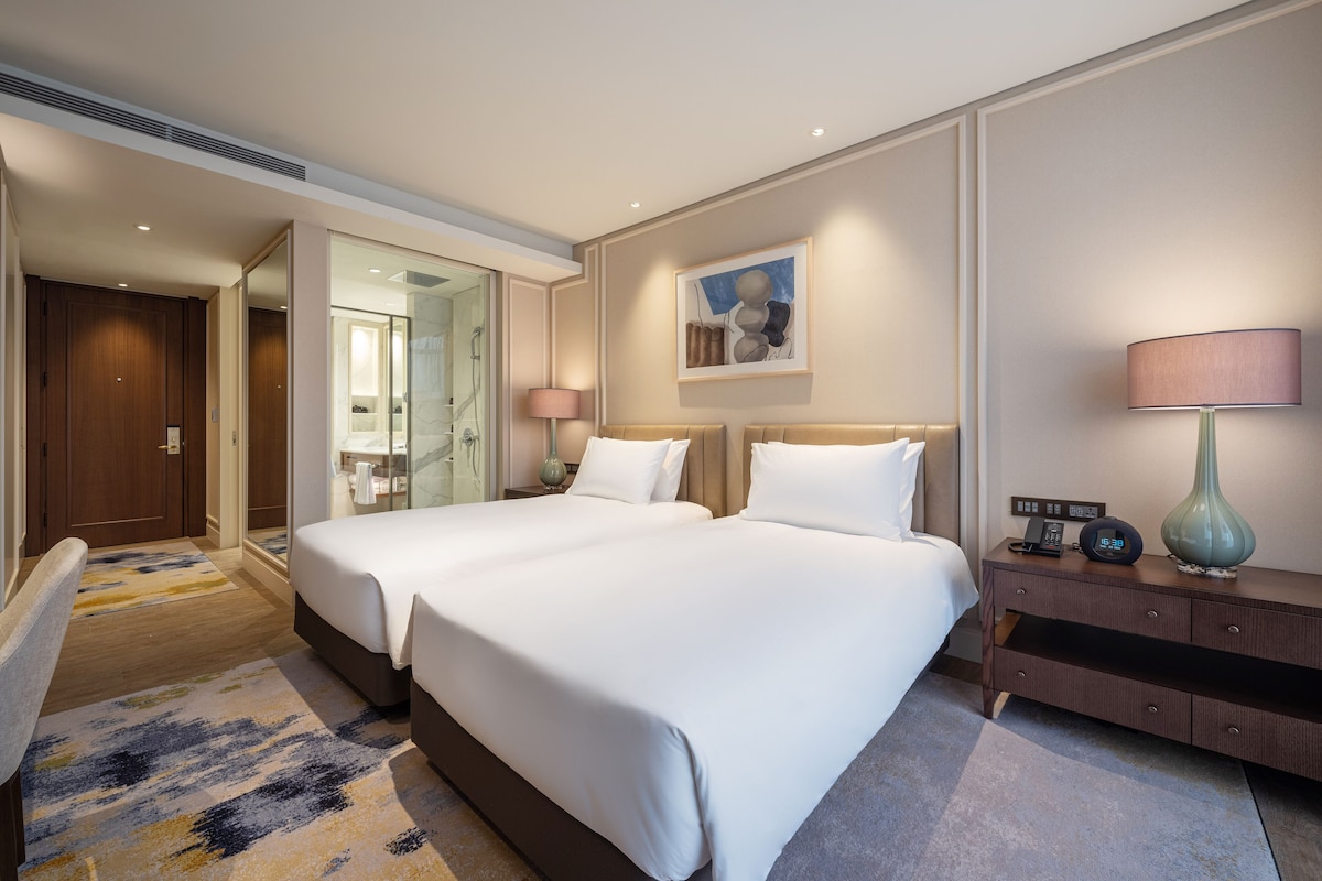 A hotel room with two beds, a bedside table with a lamp, a painting on the wall, and a bathroom visible through an open door.