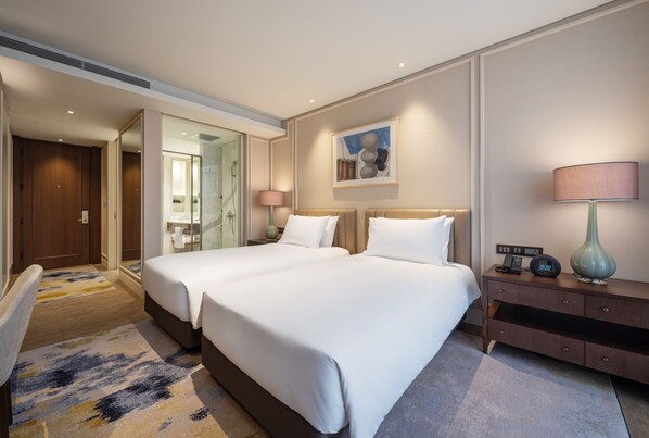 Habitación de hotel con dos camas, una mesita de noche con lámpara, un cuadro en la pared y un baño visible a través de una puerta abierta.