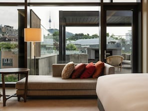Quarto de hotel com sofá, uma pequena mesa, abajur e vista para a paisagem urbana.