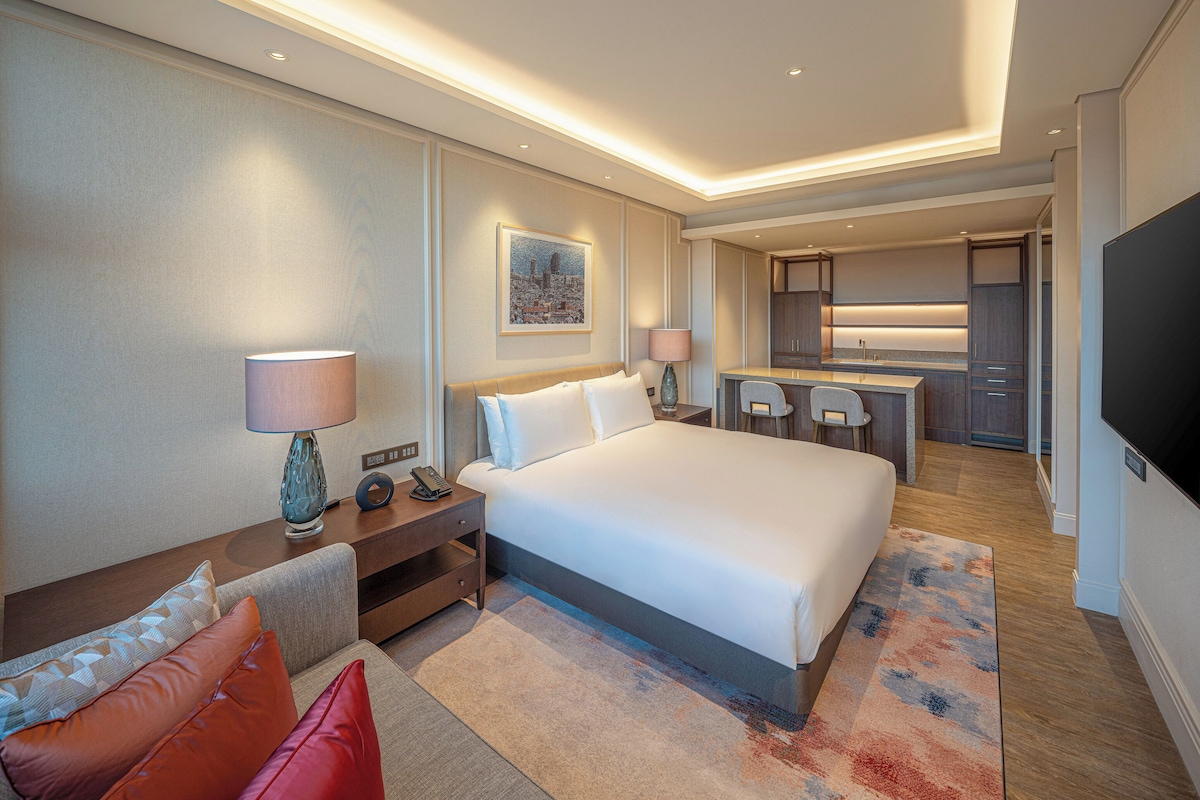 A hotel room with a large bed, a desk with a lamp, a small kitchen area, and a TV.
