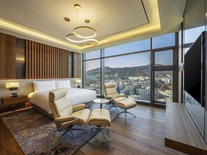 A modern hotel room with a large bed, two armchairs, a small table, a TV, and a city view through large windows.
