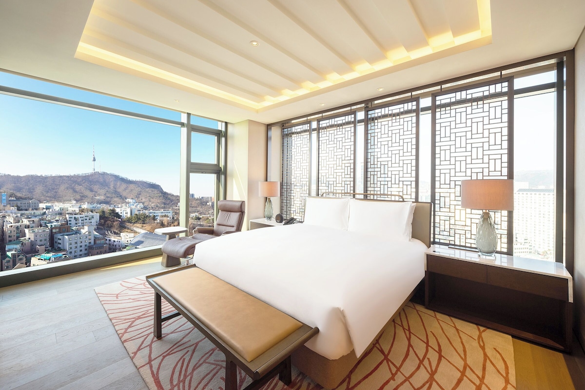 A modern hotel room with a large bed, a bench, a desk, and a view of a cityscape.