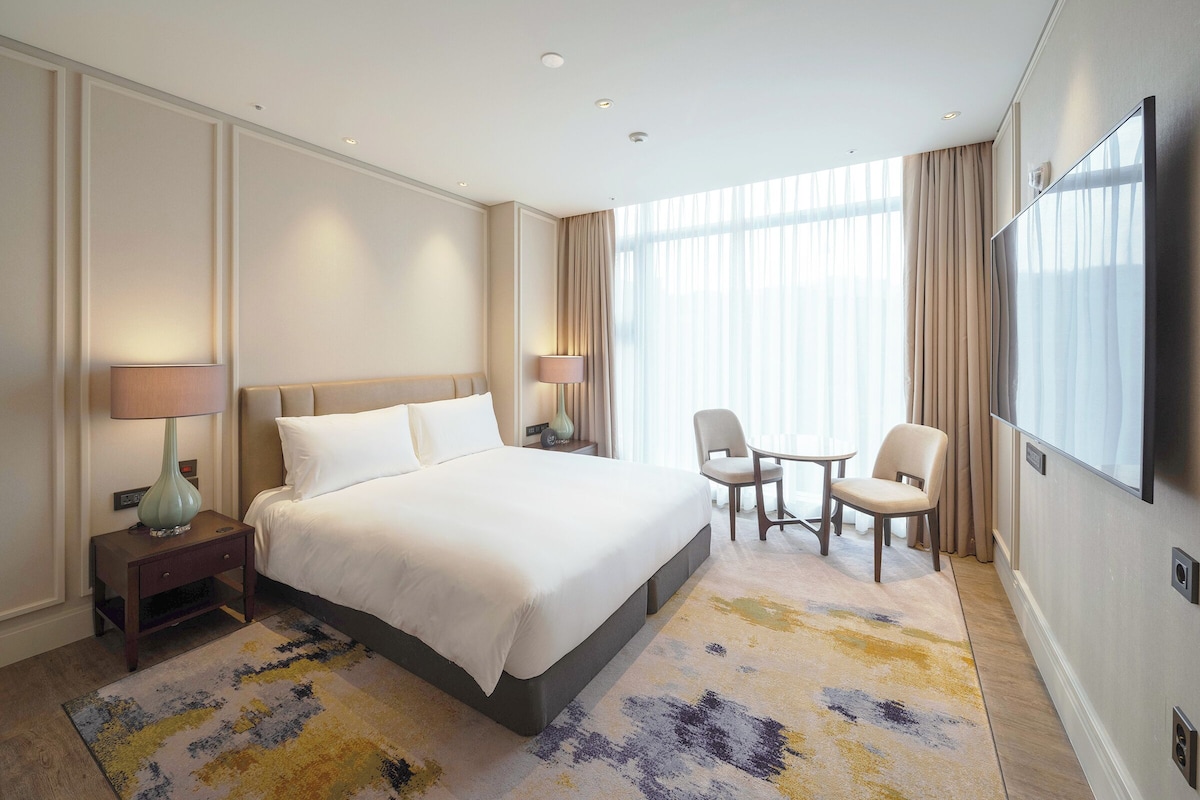 A modern hotel room with a large bed, a desk with a lamp, a small table with two chairs, a TV, and a large window with curtains.