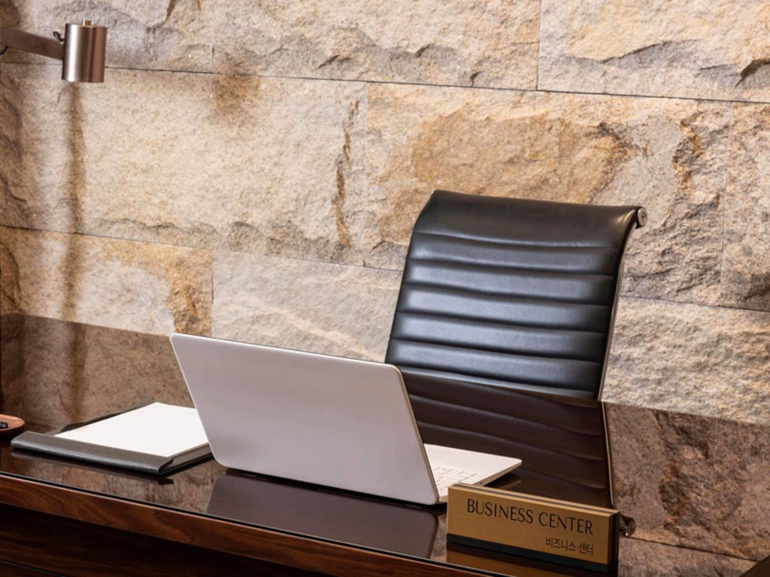 A business center with a laptop on a desk, a leather chair, and a stone wall in the background.