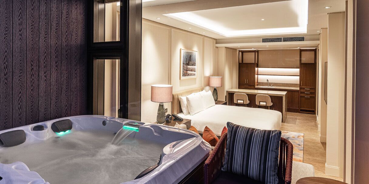 A hotel room with a large bed, a sitting area with a chair, a desk, a kitchenette, and a hot tub.