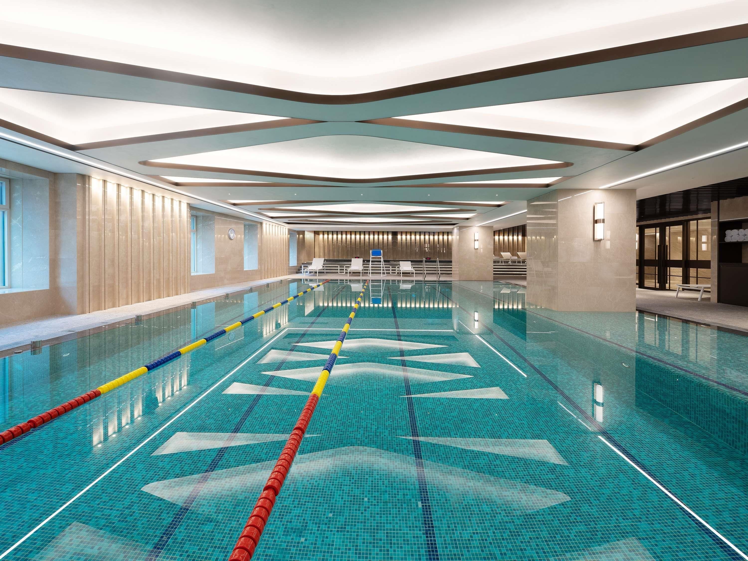 A large indoor swimming pool with clear lanes, a modern ceiling design, and a seating area with chairs.