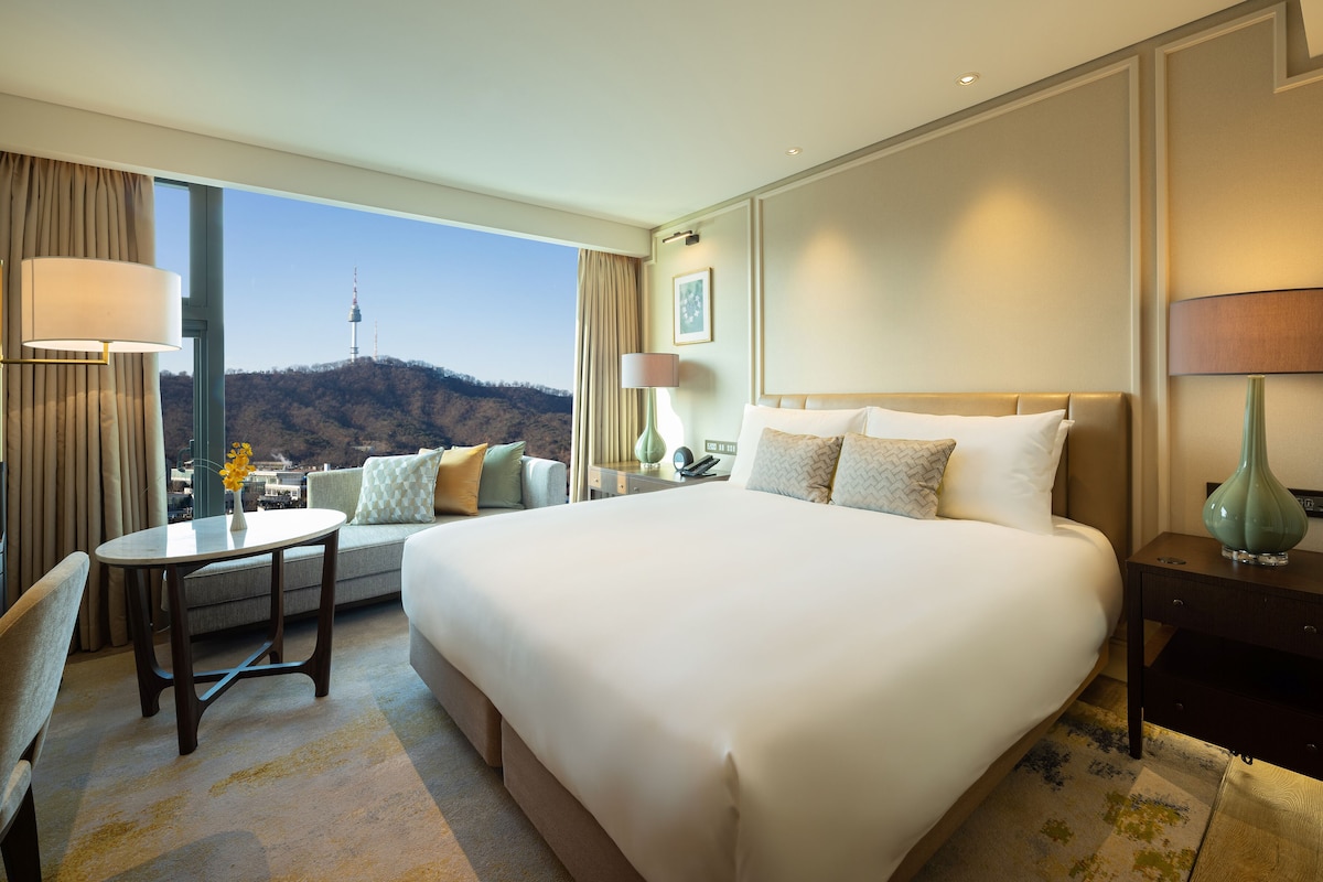 A hotel room with a large bed, a desk, a chair, a lamp, and a view of a cityscape and mountains.