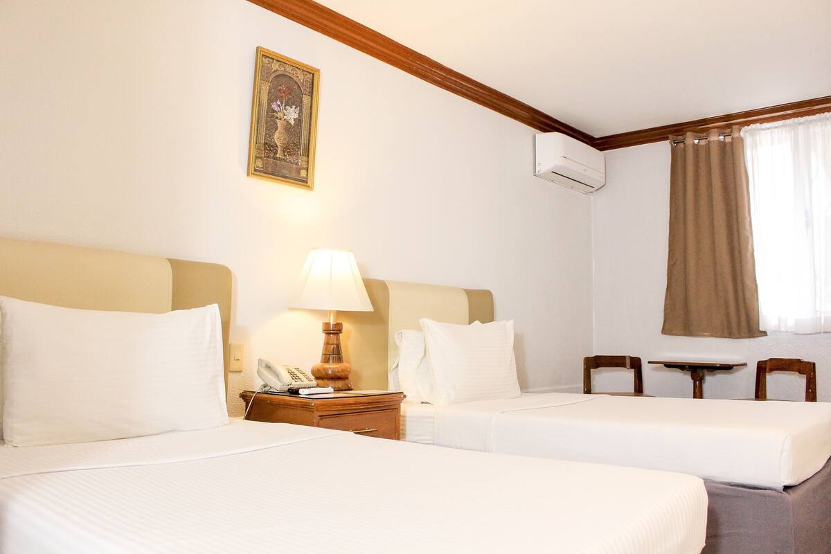 A hotel room with two beds, a nightstand with a lamp, a small table, and a chair.