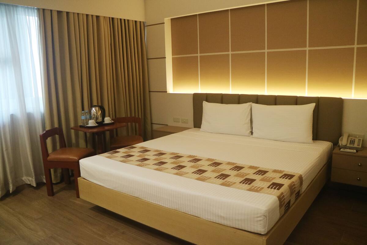 A hotel room with a large bed, a bedside table with a lamp, a chair, and a window with curtains.