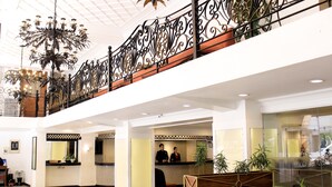 Un hall d’hôtel comprenant un comptoir d’accueil, un escalier orné de balustrades travaillées et un lustre.