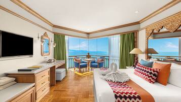 A hotel room with a large bed, a desk, a TV, a balcony with a view of the sea, and a dining area with chairs.