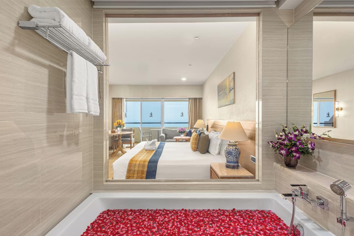 Ett hotellrum med ett stort fönster, en säng med ett blå- och gulrandigt överkast, ett sängbord med en lampa och ett badrum med ett badkar fyllt med röda rosenblad.