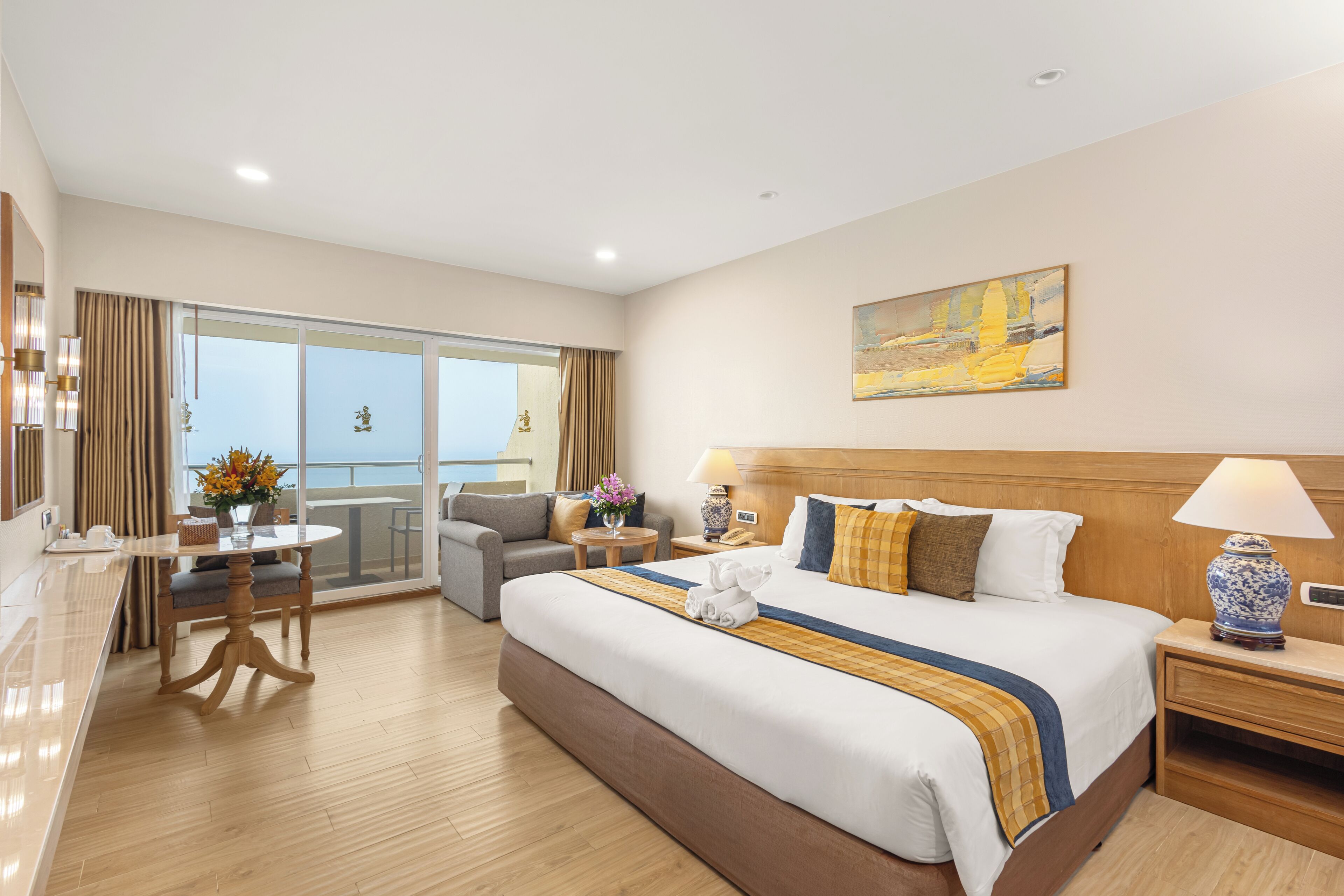 A hotel room with a large bed, a sitting area with a table and chairs, a balcony with a view, and a painting on the wall.