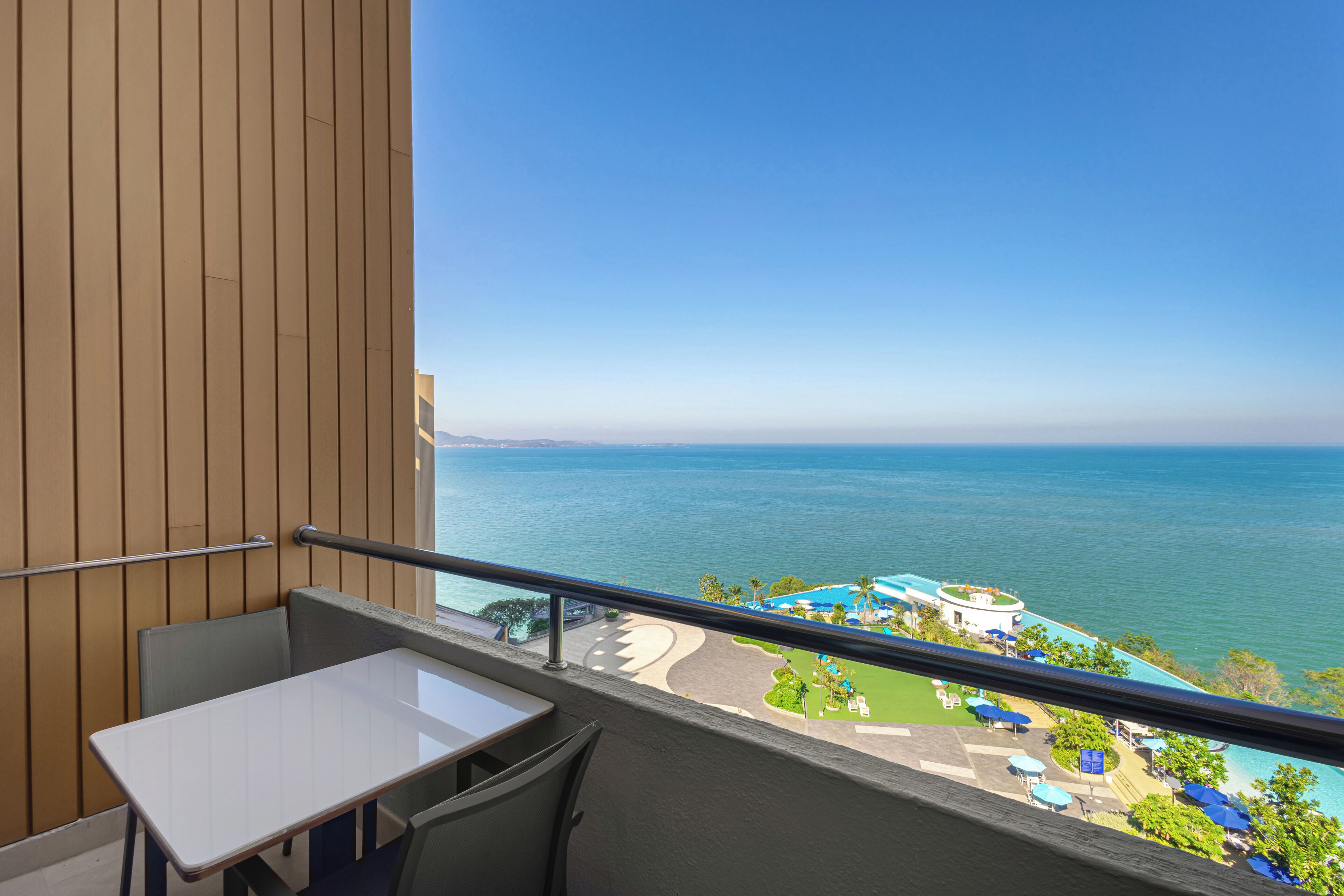 A balcony with a table and chairs, overlooking a pool and resort area with a clear view of the sea.