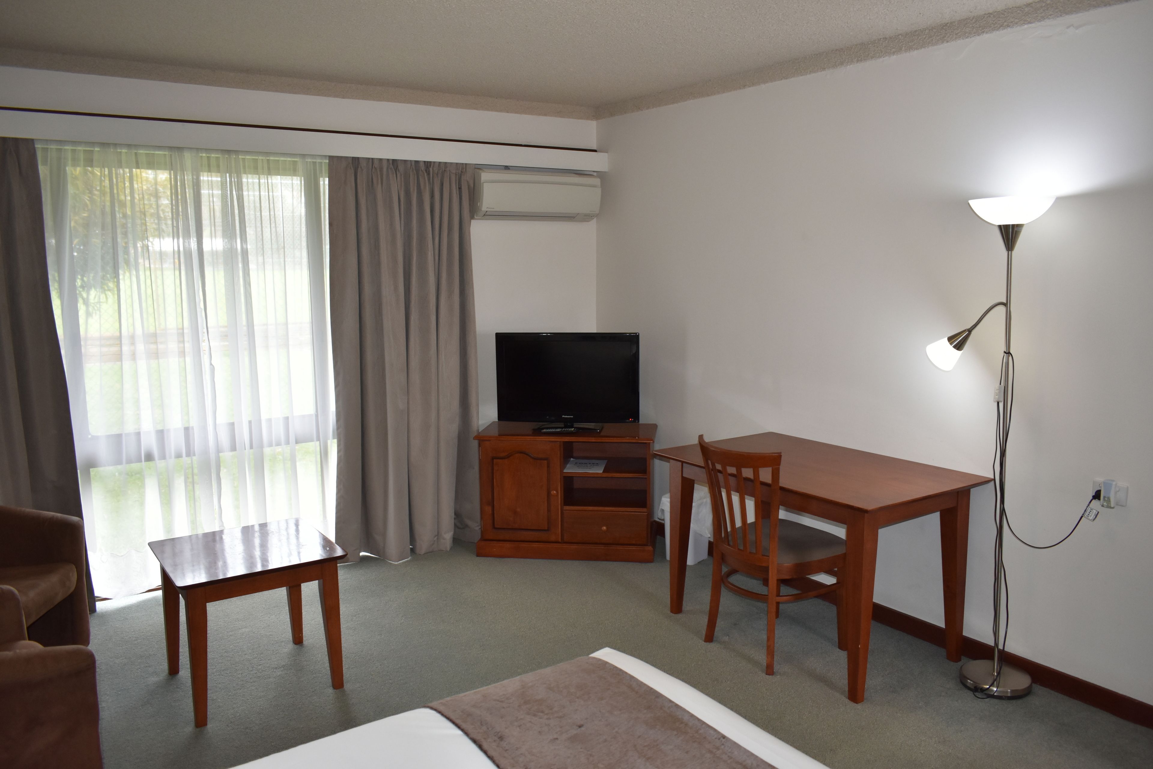 Una habitación de hotel con una cama, un escritorio de madera, un televisor y una lámpara de pie.