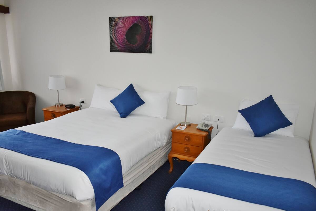 Habitación de hotel con dos camas, cada una con cubrecama azul y blanco, un sillón marrón, una mesita de noche con lámpara, y una obra de arte colgada en la pared.