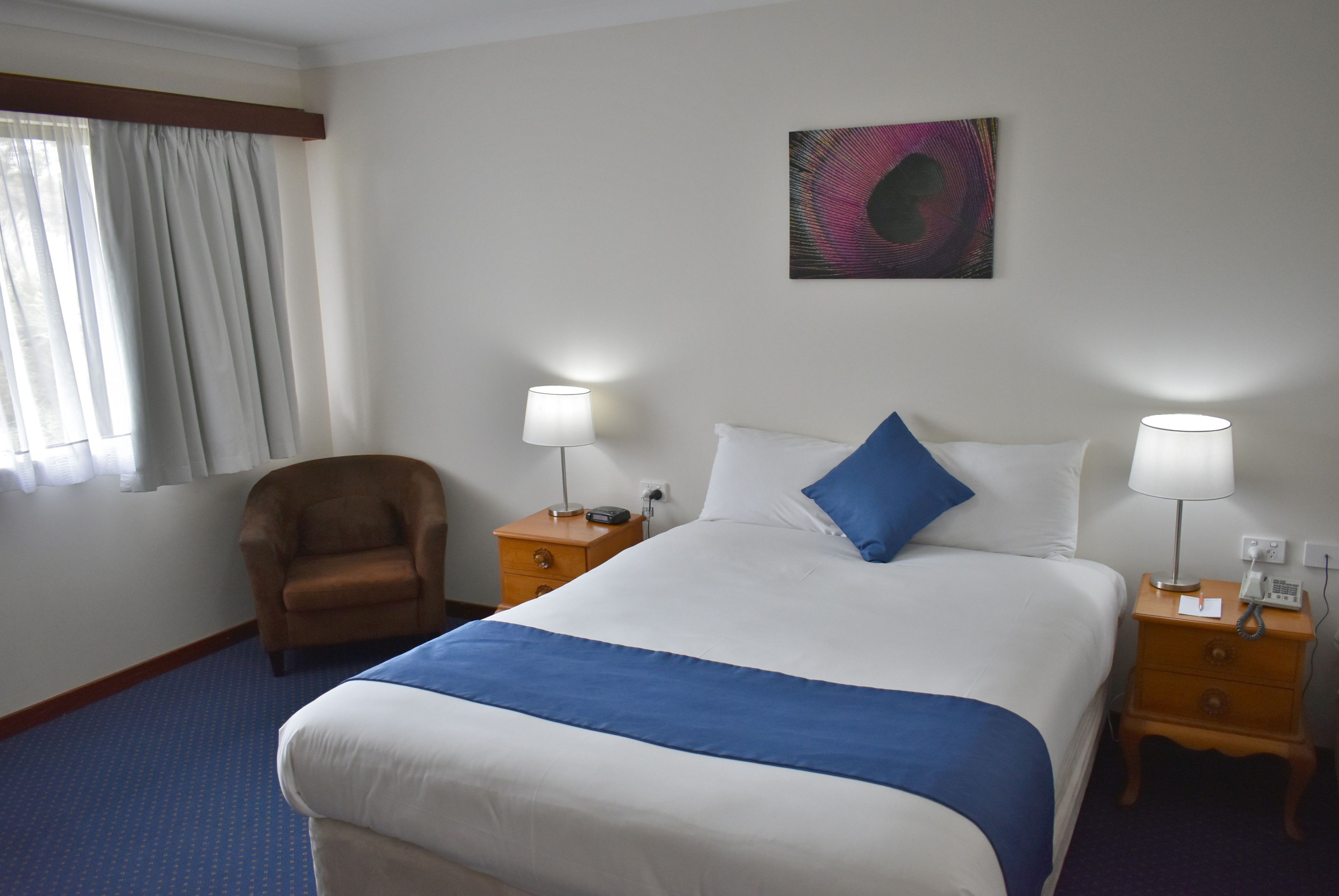 Una habitación de hotel con una cama grande, dos mesitas de noche con lámparas, una silla, un ventanal con cortinas y una obra de arte colgada en la pared.