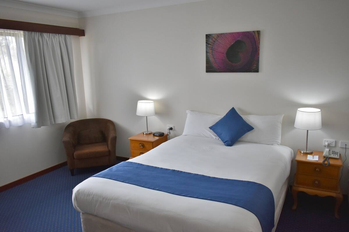 Una habitación de hotel con una cama grande, dos mesitas de noche con lámparas, una silla, un ventanal con cortinas y una obra de arte colgada en la pared.