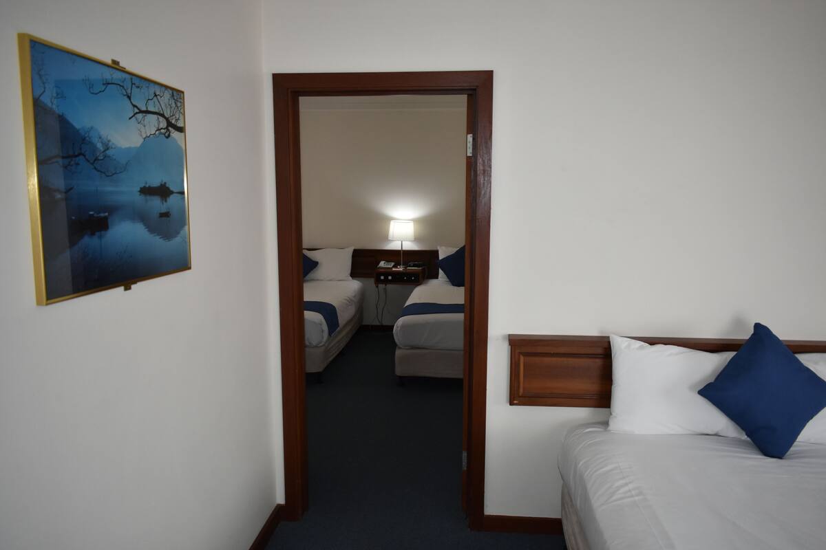 Habitación de hotel con cama, mesita de noche, lámpara, un cuadro y vista a otra habitación con dos camas.
