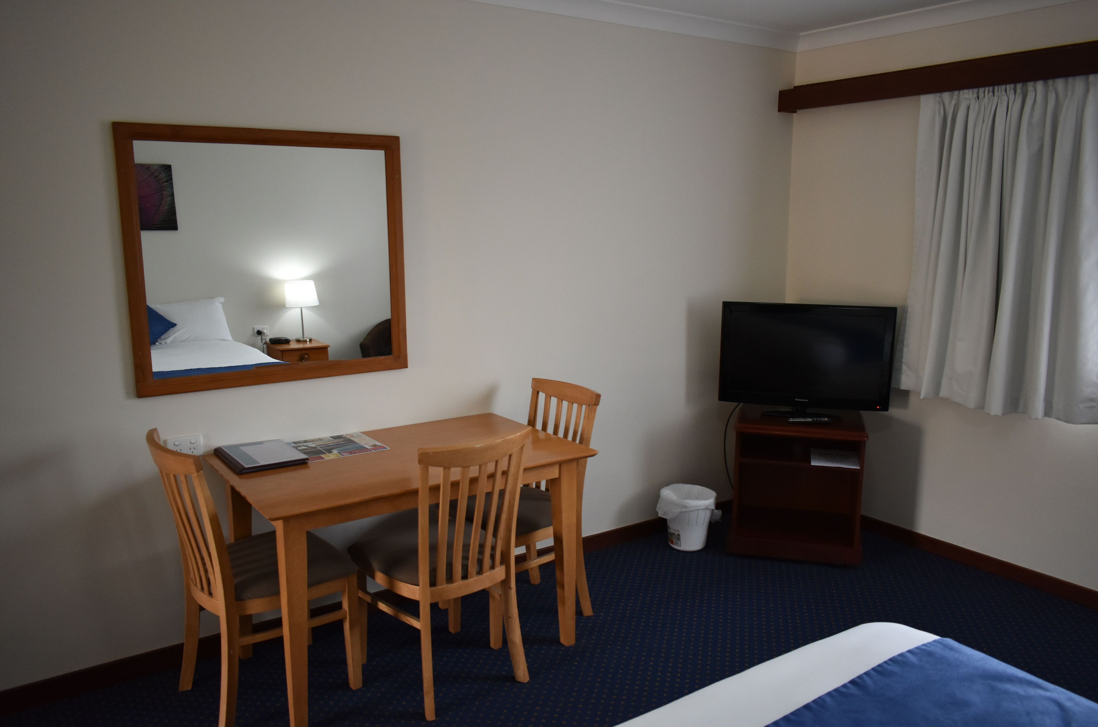 A hotel room with a bed, a TV, a desk with chairs, a mirror, and a small trash bin.
