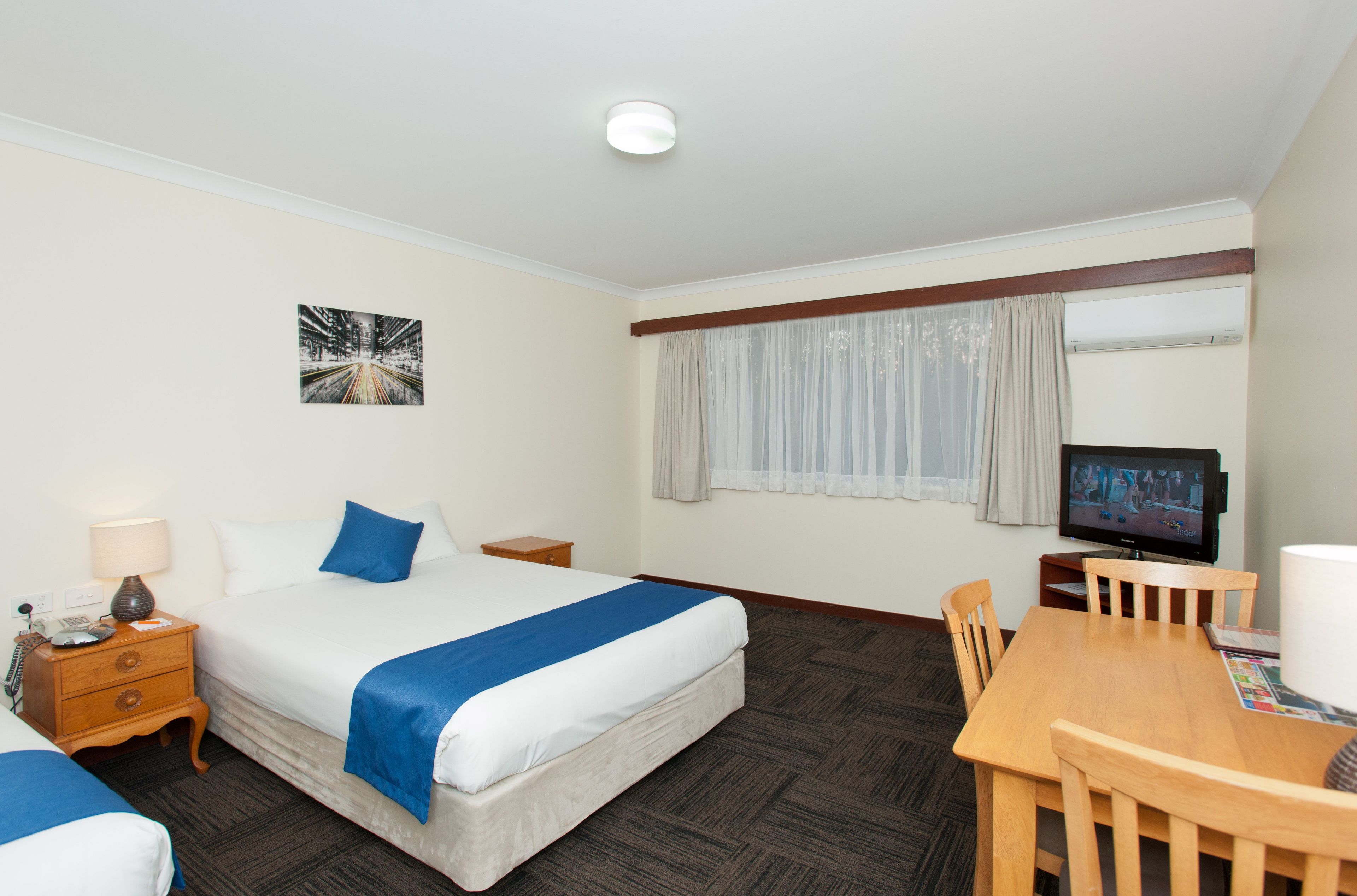 Een hotelkamer met een bed, nachtkastje, lamp, televisie en een eettafel met stoelen.
