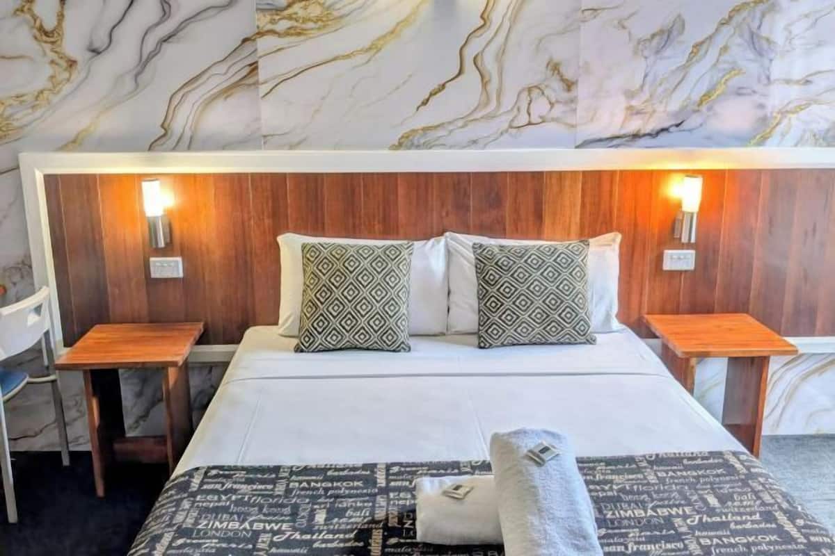 A hotel room with a marble-patterned wall, a wooden headboard, two bedside tables with lamps, a bed with patterned pillows, and a blanket with city names.