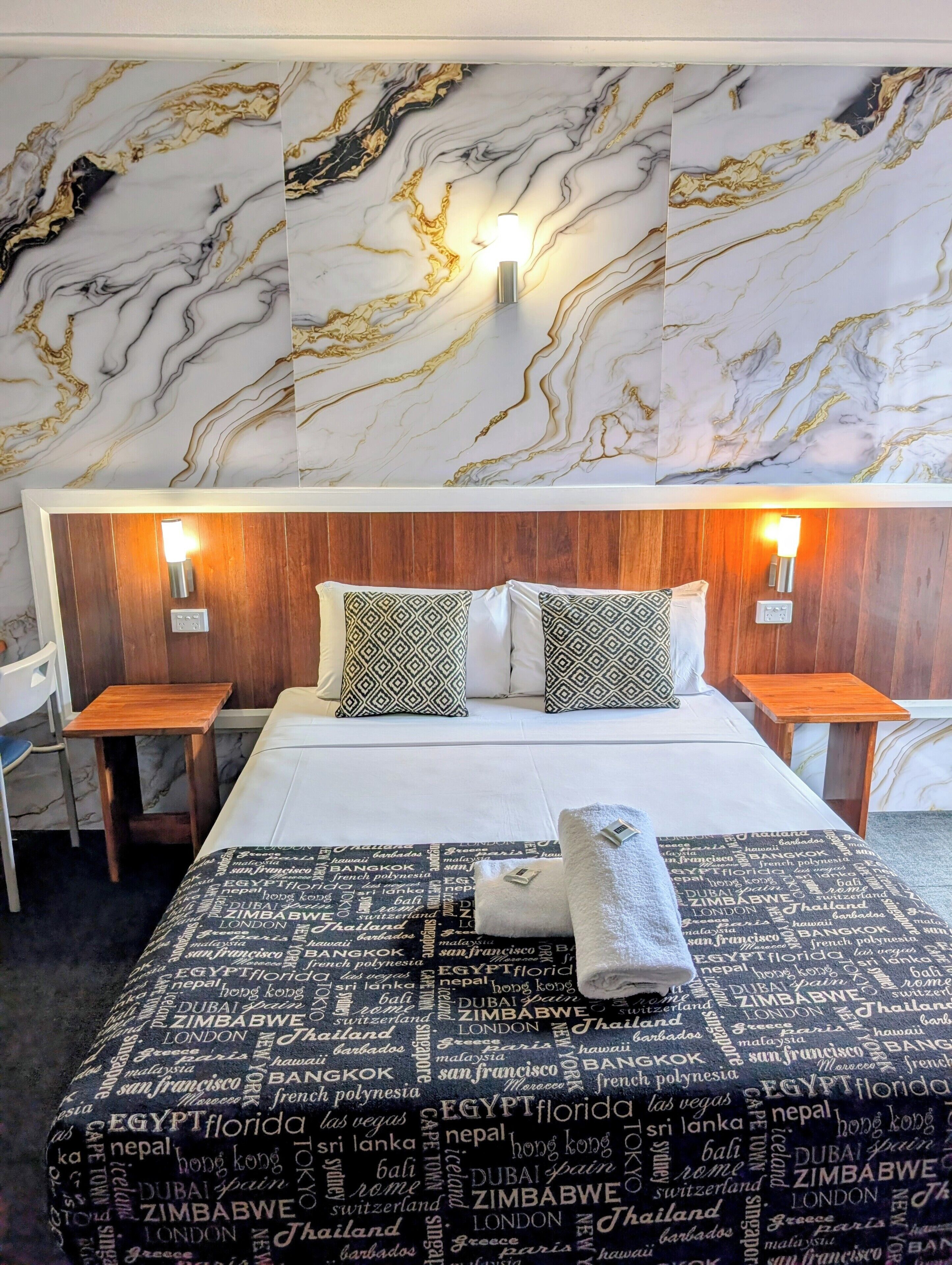 A hotel room with a marble-patterned wall, a neatly made bed with patterned pillows, two bedside tables, and a wall-mounted light fixture.